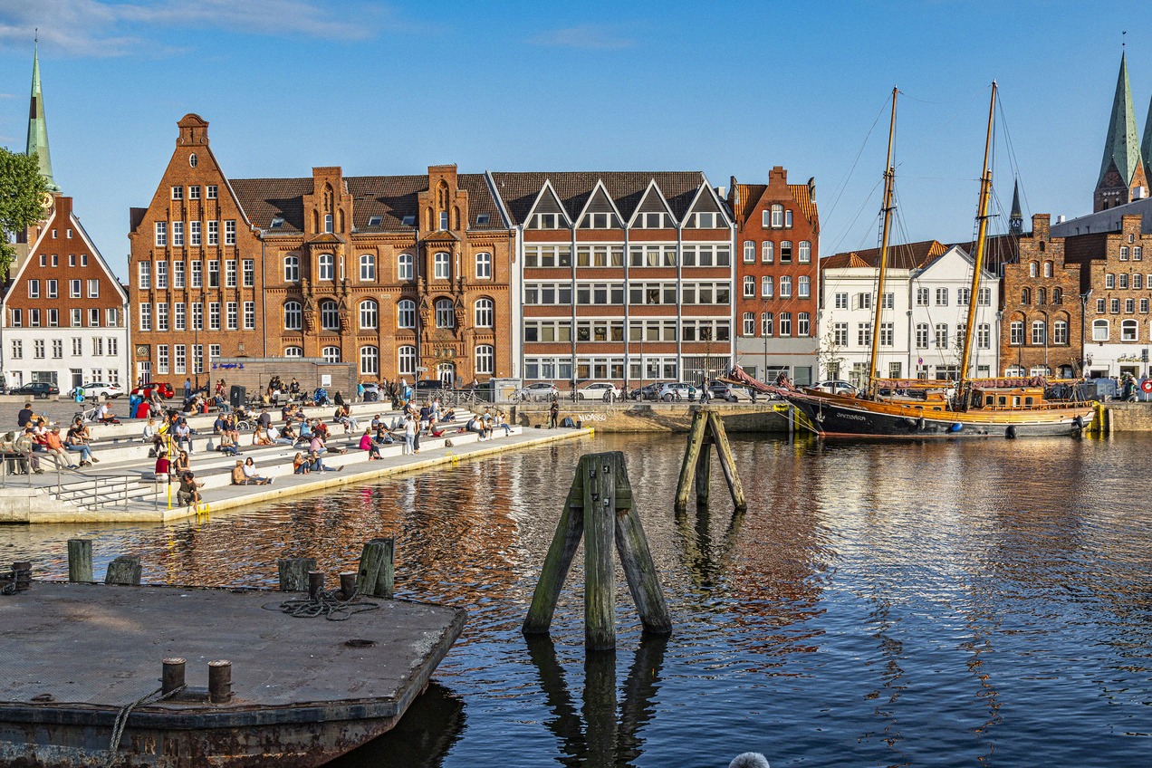 Belebter Kai an der Trave in Lübeck mit historischen Backsteinlagerhäusern, einem alten Segelschiff und entspannenden Menschen in der Abendsonne